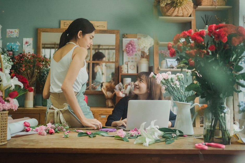 A florist and a customer collaborate inside a brightly lit flower shop, with the florist standing and wearing a beige apron and white sleeveless top, while the customer, seated at a wooden table, has 