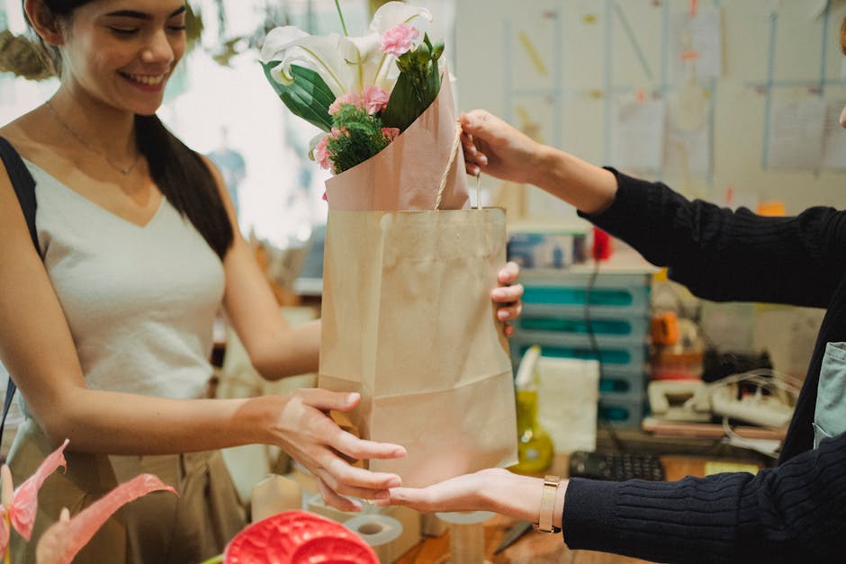 A woman with long dark hair, wearing a white sleeveless top, is smiling while handing over a bouquet of fresh flowers wrapped in pink and brown paper. The bouquet contains white lilies, pink roses, an