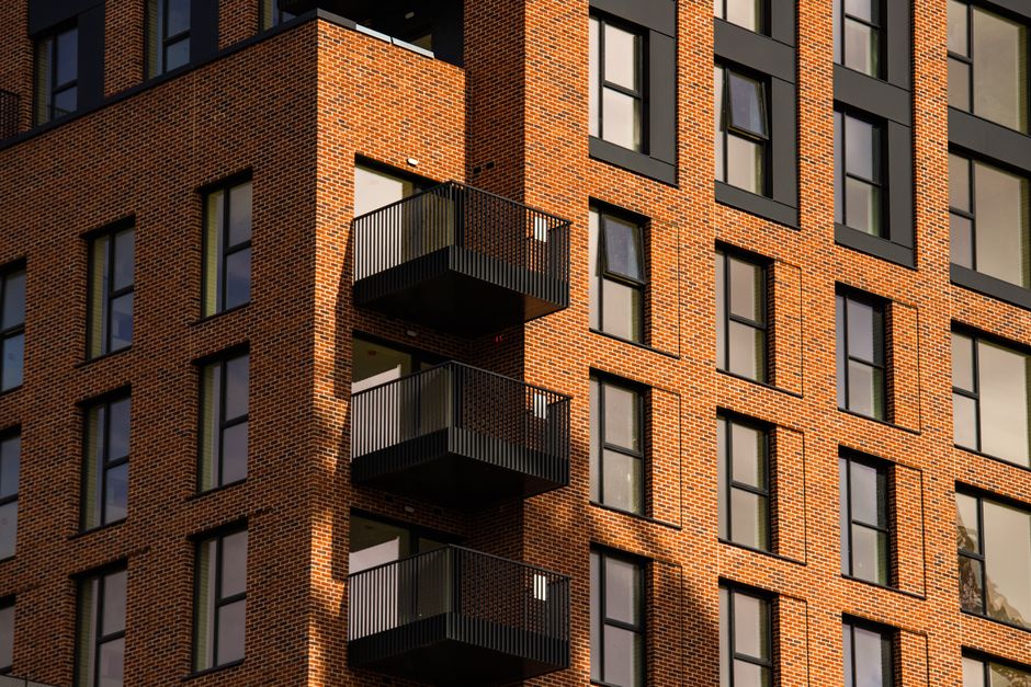 A close-up view of a modern multi-story brick residential building with several black metal balconies. The building features a grid pattern of rectangular windows with black frames, interspersed with 