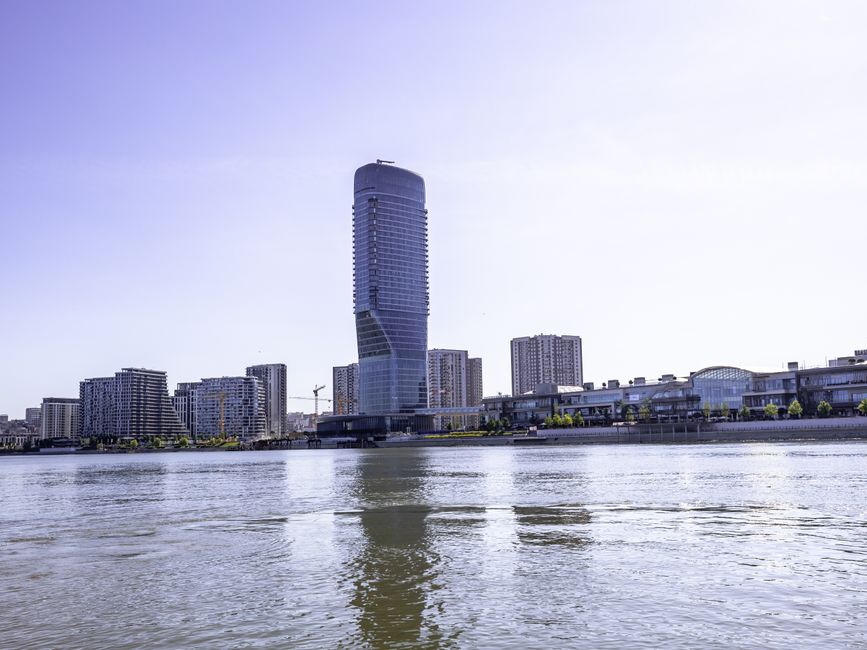 A modern cityscape featuring a tall, curved skyscraper with a glass facade, surrounded by shorter commercial and residential buildings along a riverbank, with reflections of the buildings visible in t