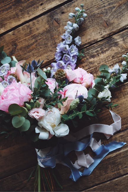A bridal floral bouquet arranged with large, soft pink peonies and white roses, accented by sprigs of eucalyptus leaves and small, white filler flowers, with some dark blue thistle-like blooms adding 