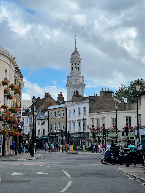A lively street scene in Kidbrooke featuring a central clock tower with a tall, ornate spire topped by a golden weather vane, surrounded by historic-style buildings with white and beige facades, some 