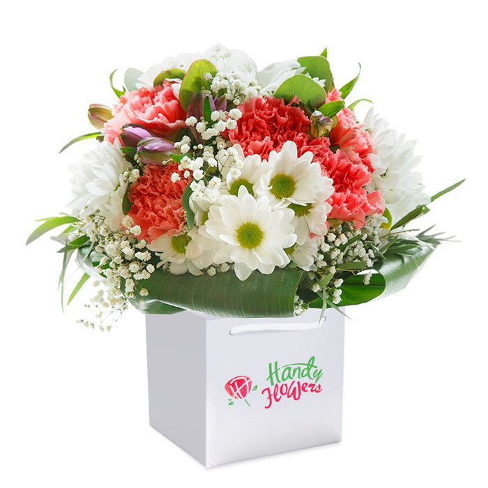 Poised elegantly within a brilliant white square gift bag, this lush bouquet exudes the vibrancy and artistry synonymous with Kidbrooke's professional florists. Plush coral-pink and vivid red carnations take centre stage, each flower boasting a profusion of delicately ruffled petals that speak to freshness and attention to detail. Threaded among them are pristine white daisies, each with a distinct lime-green core, bringing a spark of cheerful contrast and echoing the lively energy found near Kidbrooke's greenways. Baby's breath, with its minuscule white clusters, imparts a soft, cloud-like texture, artfully filling gaps and adding breathability to the design. On the left edge, a few closed buds-touched with mauve and purple-interject a gentle mystery, hinting at blooms yet to reveal their charm. Surrounding the bouquet, deep green foliage fans out in smooth arcs, their broad, glossy leaves creating a serene, natural frame-the type of expertly arranged greenery that makes each bouquet from a local Kidbrooke florist memorable. The pure, neutral backdrop ensures every nuance is visible and celebrated, from the texture of a carnation's ruffle to the crisp shine on a leaf. Ideal for life's joyful occasions-birthdays, new beginnings, heartfelt thanks-this detailed arrangement reflects both skilled craftsmanship and the joy of giving or receiving fresh, locally delivered flowers in Kidbrooke.