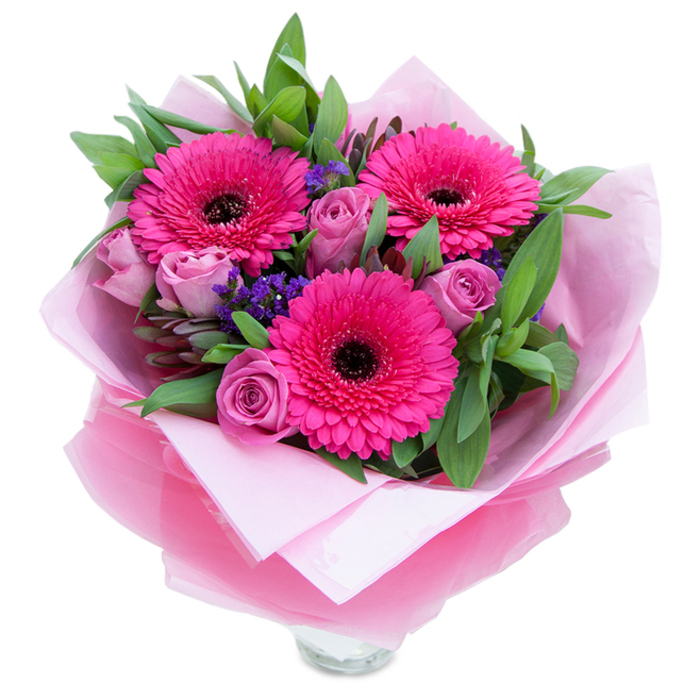I see this hand-tied bunch as a gentle celebration of springtime colour, centred on three bold fuchsia Gerbera daisies whose many petals radiate outward like little fireworks. Their dark centres anchor the composition while softer lavender-pink roses, in a muted dusty-rose tone, unfurl nearby, their tight spirals promising a quiet, powdery fragrance. Interspersed throughout are clusters of purple statice - tiny, papery florets that create a pleasing, textural punctuation against the smoother rose petals and broad leaves. The greenery, likely salal or eucalyptus, offers a cool, dewy green that supports the palette and adds a slightly aromatic, almost menthol note when the bouquet is handled. The stems are wrapped with care: a pale pink matte tissue forms a gentle cradle, then a translucent fuchsia-pink cellophane, slightly crinkled, envelopes the blooms with a soft shimmer that catches the eye without overpowering the flowers. You can glimpse the top rim of a clear glass vase at the base, suggesting practical readiness for immediate display. Photographed in soft daylight on a plain white surface, the arrangement conveys intimacy and attentiveness - the kind of gift to lift spirits on a bedside table, to mark a thoughtful anniversary, or to brighten a family gathering in Kidbrooke Village or an afternoon picnic at Sutcliffe Park. The bouquet reads as both playful and refined, handmade with a florist's attention to balance, scent and texture.