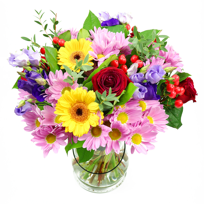 A full, dome-shaped bouquet overflows with mixed flowers, rising from a simple clear glass vase and set against a smooth white background that accentuates every tone and texture. At the heart of the arrangement sit deep red roses, their velvety petals layered in tight, romantic spirals that give the design depth and elegance. Nearby, two bold yellow gerbera daisies with rich brown-orange centres add a burst of brightness, their wide petals forming cheerful circles of colour. Surrounding these focal blooms is a soft halo of pink daisies or chrysanthemums, each flower composed of numerous slender petals and a tiny yellow centre, lending the bouquet a plush, cushion-like fullness and a reassuring, gentle charm. Woven throughout are slender stems of lisianthus in purple and lavender hues, their bell-shaped blossoms and ruffled edges introducing cool, sophisticated notes that balance the warmth of the reds and yellows, much like the contrast between Kidbrooke's modern developments and its green open spaces. Nestled among the petals, clusters of round, glossy red hypericum berries provide eye-catching texture and a contemporary twist. A variety of lush green foliage-broad, dark leaves and more delicate, lighter sprigs-frames the flowers, adding depth, movement, and a natural, just-arranged feel. Through the transparent vase, the neatly cut stems are clearly visible in their bath of clear water, reinforcing the sense of freshness and skilled floristry that you might expect from a trusted Kidbrooke flower specialist. The overall bouquet radiates joy and considered beauty, ideal for celebrations, get-well wishes, or welcoming someone to a new home in the Kidbrooke area.