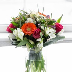 Rich in colour and spirited in design, this masterful bouquet draws inspiration from the vibrancy of Kidbrooke's local gardens. Soft peach gerberas, their petals gently ruffled and warming the scene, are embraced by brilliant orange counterparts that radiate cheer. Velvet magenta roses nestle between the lighter tones, infusing the arrangement with a deep sense of romance and sophistication. Around and within, snowy white alstroemeria and petite blossoms flicker with brightness. The lush green structure at the base, composed of wide, paddle-shaped aspidistra leaves, wraps protectively around the bouquet-much like the comforting green spaces throughout Kidbrooke. Every detail is illuminated by a diffused white light, celebrating the unique architecture and colour balance. The bouquet evokes feelings of fresh beginnings and heartfelt congratulations, perfect for everything from life's joyful milestones to casual moments of appreciation among local friends and families.